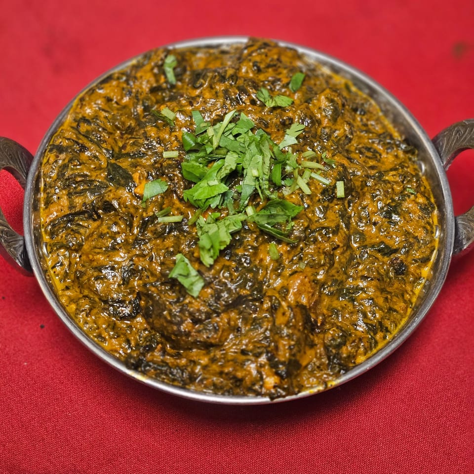 A metal bowl of Kripa Indian Cuisine's Palak Paneer, garnished with fresh cilantro, on a red tablecloth.