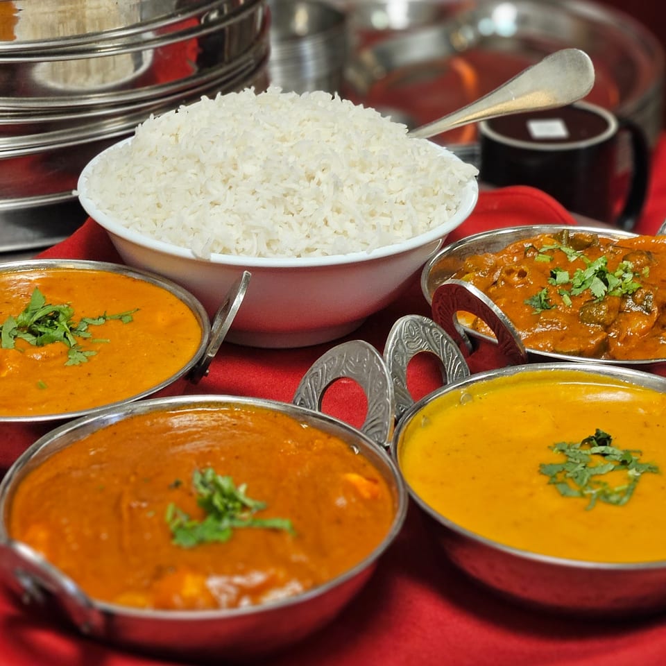 Four bowls of Kripa Indian Cuisine curries and a large bowl of white rice on a red tablecloth.